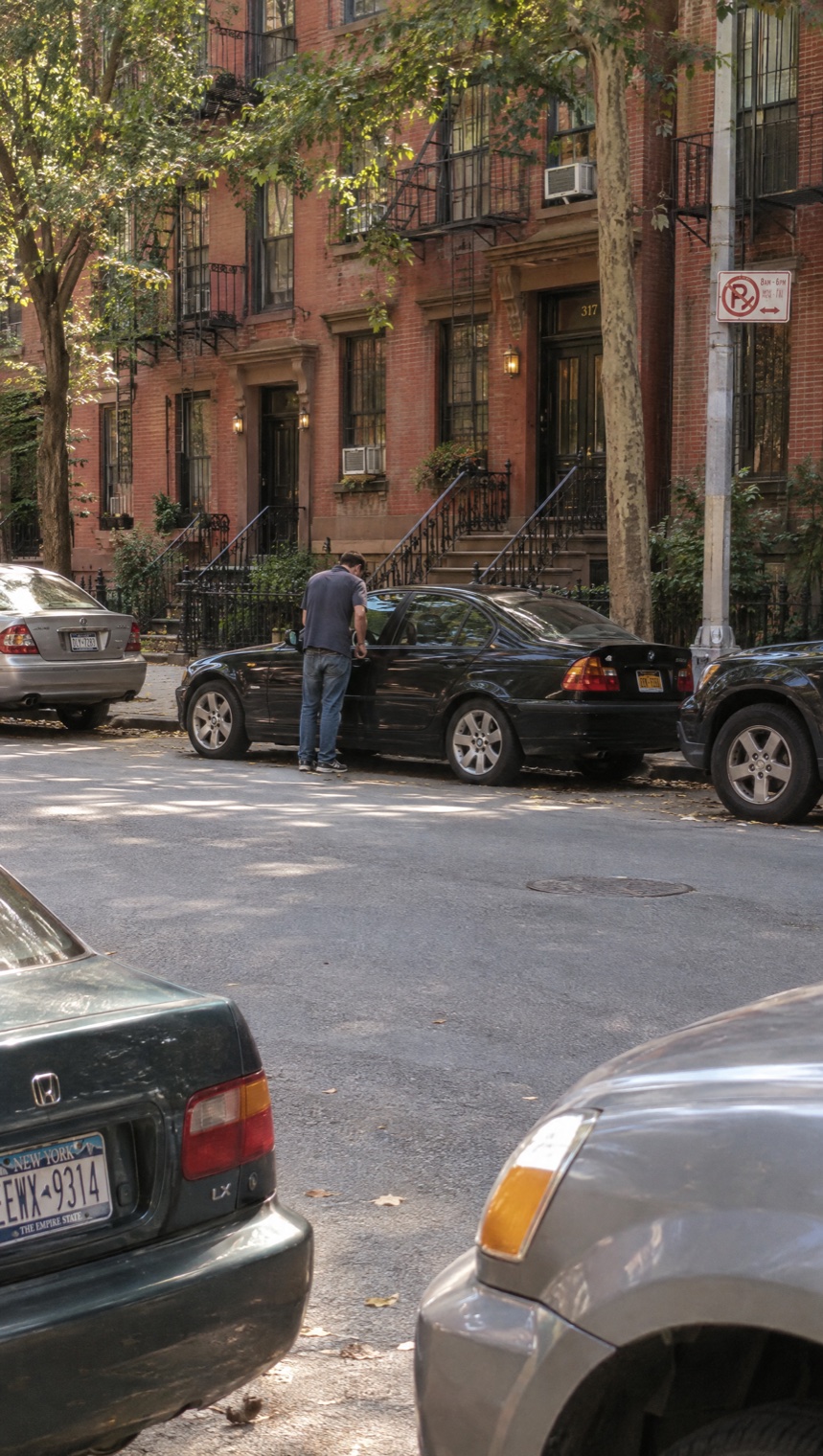 A man pulling on the door handle of a black sedan across a quiet Manhattan street, framed by two parked cars in the foreground